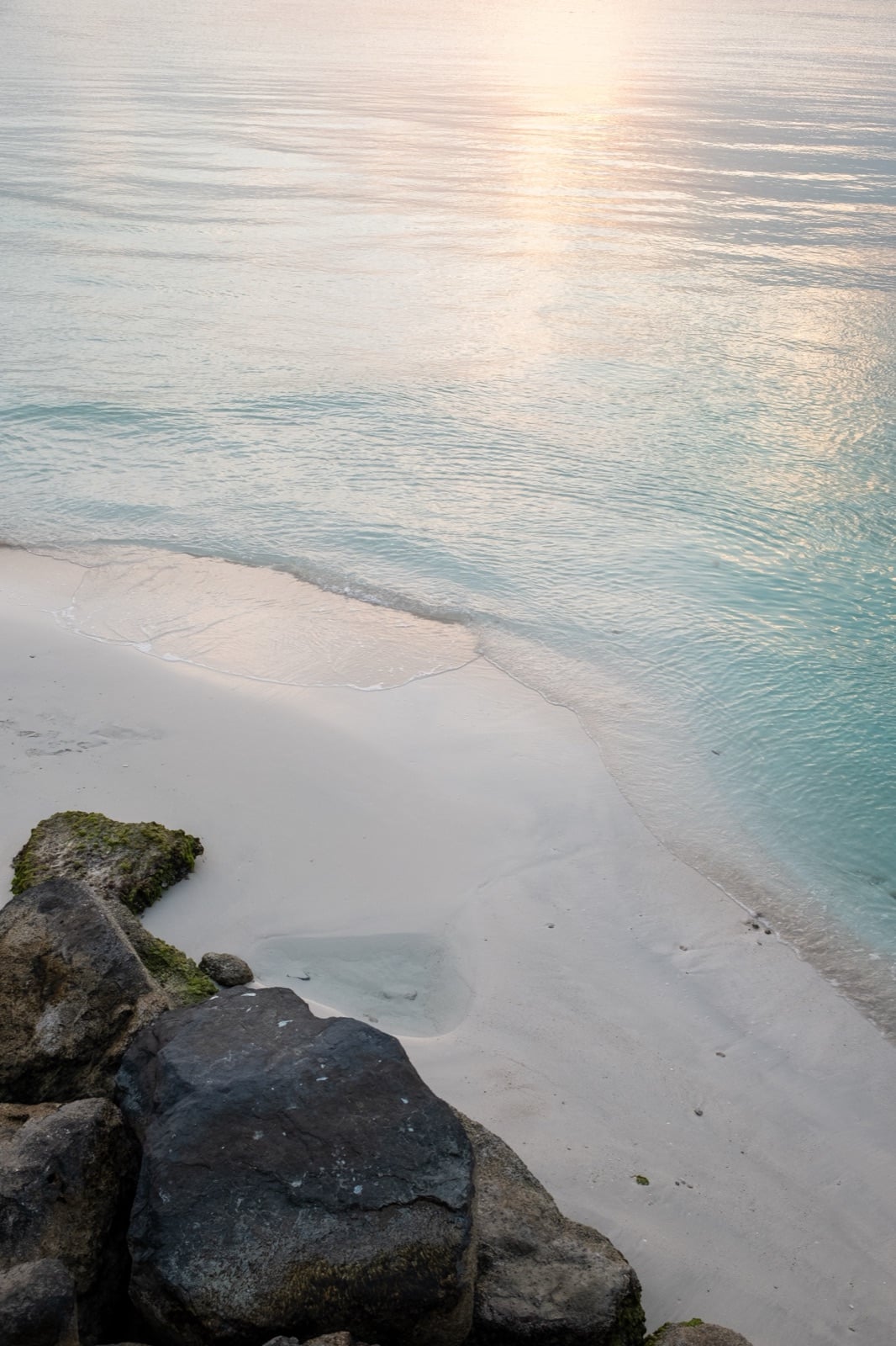 Soft sunset light on calm turquoise water, a pale beach and dark rocks in the corner